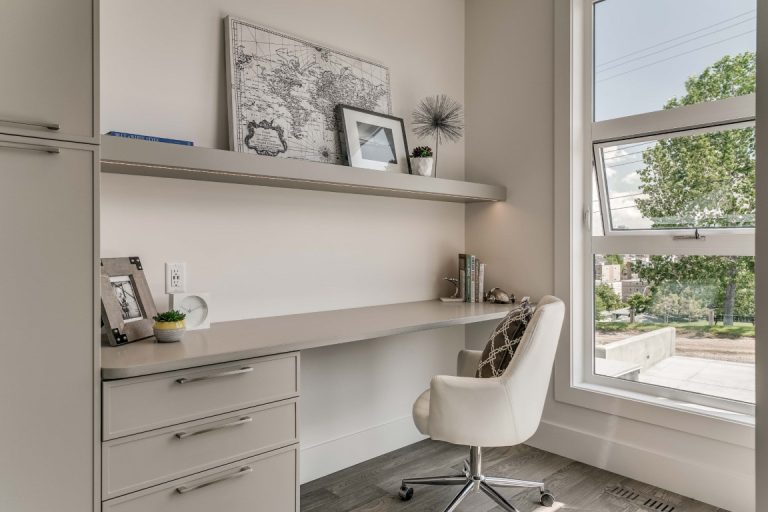Modern home office with built-in desk, storage drawers, floating shelves, white chair, and large window with natural light
